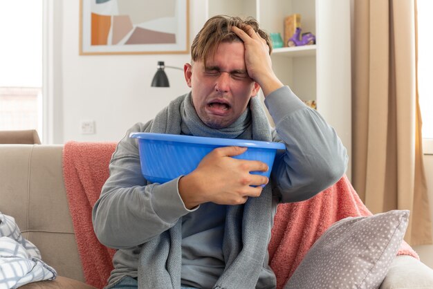 schmerzender junger kranker Mann mit Schal um den Hals, der die Hand auf seine Stirn legt und den Eimer auf der Couch im Wohnzimmer hält