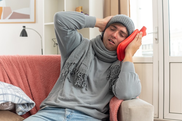 schmerzender junger kranker Mann mit Schal um den Hals, der eine Wintermütze trägt, die Hand auf den Kopf legt und eine Wärmflasche auf der Couch im Wohnzimmer hält