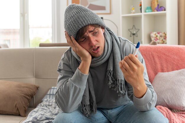 schmerzender junger kranker Mann mit Schal um den Hals mit Wintermütze, der die Hand auf sein Gesicht legt und das Thermometer auf der Couch im Wohnzimmer hält und betrachtet