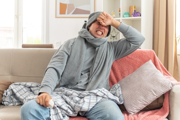 Schmerzender junger kranker slawischer Mann mit Schal um den Hals, der eine Wintermütze trägt, die Hand auf die Stirn legt und ein Taschentuch auf der Couch im Wohnzimmer hält