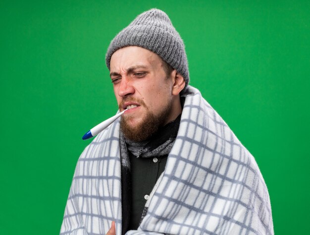 Schmerzender junger kranker slawischer Mann mit Schal um den Hals, eingewickelt in karierte Wintermütze mit Thermometer im Mund