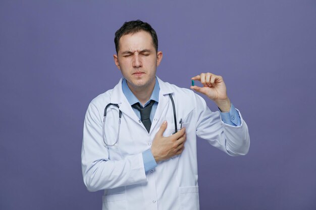 Schmerzender junger männlicher Arzt mit medizinischem Gewand und Stethoskop um den Hals, der eine Kapsel mit geschlossenen Augen zeigt, während er die Hand auf dem Herzen isoliert auf violettem Hintergrund hält