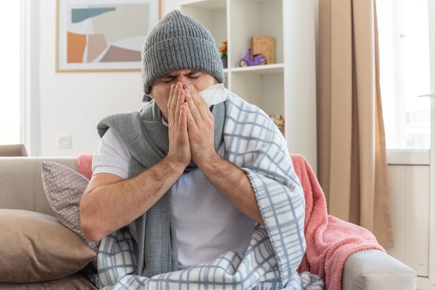 schmerzender kranker Mann mit Schal um den Hals, der eine in Plaid gewickelte Wintermütze trägt und seine Nase auf der Couch im Wohnzimmer hält