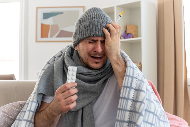 schmerzender kranker Mann mit Schal um den Hals, der eine Wintermütze trägt, die in Plaid gewickelt ist und seine Hand auf den Kopf legt, die eine Medizinblisterpackung und eine Spritze auf der Couch im Wohnzimmer hält