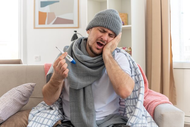 schmerzender kranker Mann mit Schal um den Hals, der eine Wintermütze trägt, die seine Hand auf das Gesicht legt und das Thermometer auf der Couch im Wohnzimmer hält