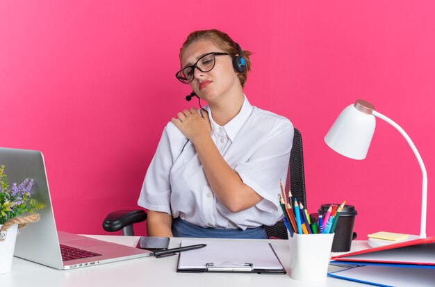 Schmerzendes junges blondes Callcenter-Mädchen mit Headset und Brille, das am Schreibtisch mit Arbeitswerkzeugen sitzt und auf den Laptop schaut, der die Hand auf der Schulter isoliert auf rosa Wand hält