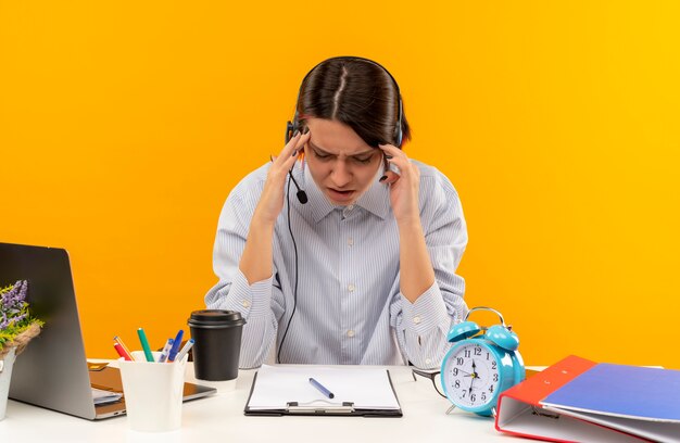 Schmerzendes junges Callcenter-Mädchen, das ein Headset trägt, das am Schreibtisch sitzt und Hände auf den Kopf legt, der unter Kopfschmerzen mit geschlossenen Augen leidet, die auf Orange isoliert werden