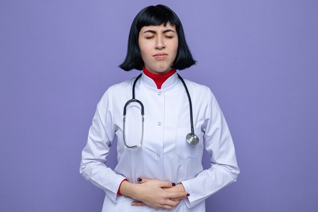Schmerzendes junges hübsches kaukasisches Mädchen in Arztuniform mit Stethoskop, das die Hände auf den Bauch legt