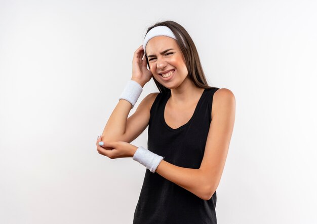 Schmerzendes junges hübsches sportliches Mädchen mit Stirnband und Armband, das die Hände auf den Arm und den Kopf auf die weiße Wand legt
