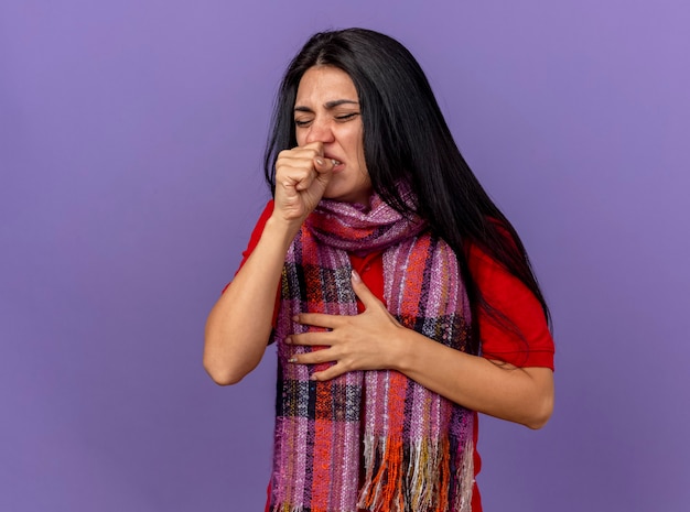Schmerzhafte junge kranke Frau, die Schal trägt Hand auf Brust mit geschlossenen Augen hustend hält Faust nahe Mund isoliert auf lila Wand