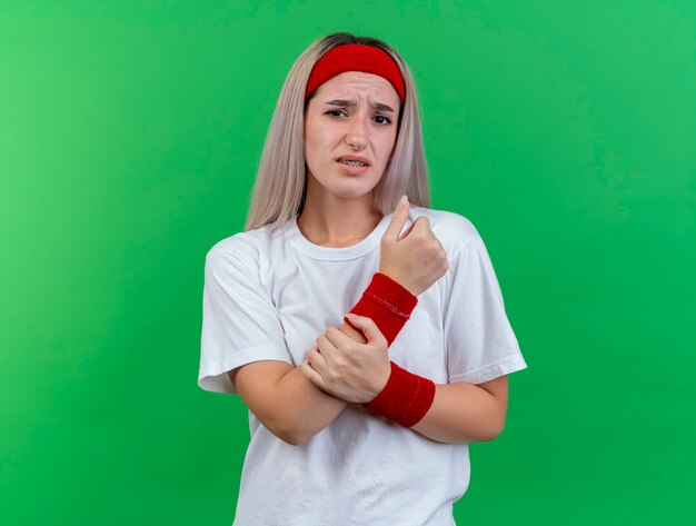 Schmerzhafte junge sportliche Frau mit Zahnspangen, die Stirnband und Armbänder tragen, hält Hand lokalisiert auf grüner Wand