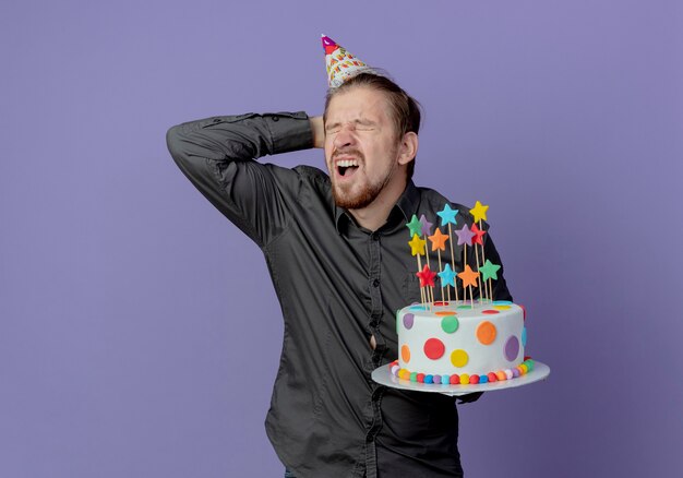 Schmerzhafter gutaussehender Mann in der Geburtstagskappe hält Kopf hinter und hält Kuchen lokalisiert auf lila Wand