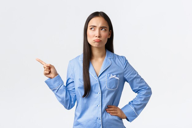 Schmollend enttäuschtes asiatisches Mädchen im blauen Pyjama, die Stirn runzelnd und schmollend, als ob es auf die obere linke Ecke zeigt, unzufrieden aussieht, mit Abneigung anstarren, sich als stehender weißer Hintergrund beschweren