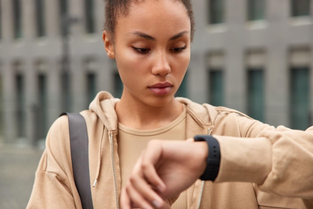 Schnappschuss einer ernsthaften sportlichen Frau in braunem Sweatshirt, die verbrannte Kalorien auf der Smartwatch am Handgelenk überprüft, hat gesunde Hautposen vor verschwommenem Hintergrund, Übungen im Freien Sportkonzept