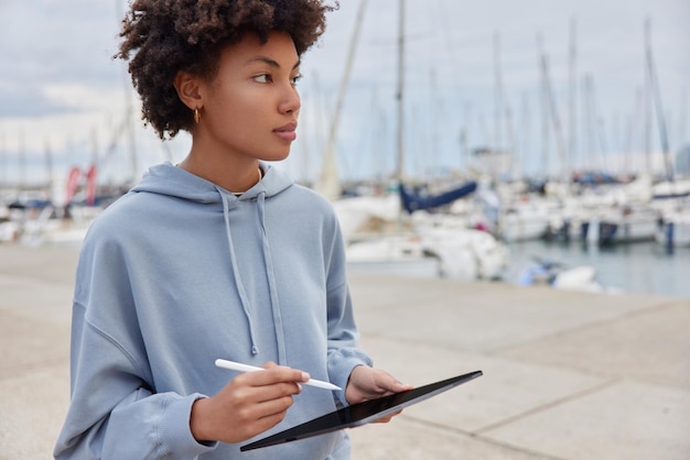 Schnappschuss einer nachdenklichen Frau macht Zeichnungen mit Tablet und Stylus blickt in die Ferne Spaziergänge im Seehafen trägt lässigen Hoodie Qualifizierte Grafikdesignerin arbeitet aus der Ferne mit Touchpad