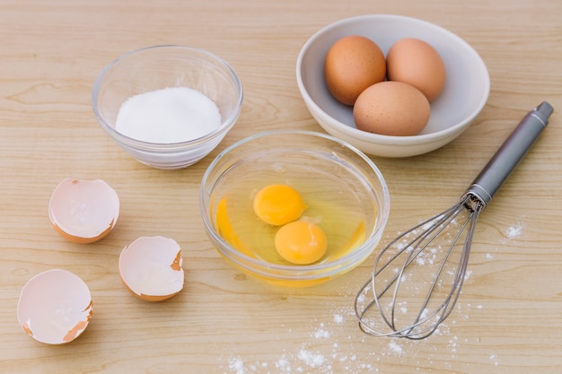 Schneebesen; Eier und Eigelb mit Muscheln und Mehl zum Backen von Kuchen auf Schreibtisch aus Holz