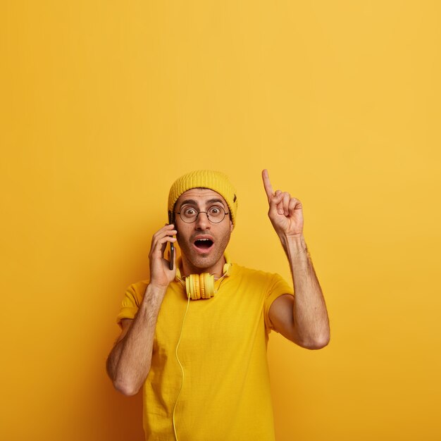 schockiert besorgter junger Mann überrascht von lästigen Neuigkeiten, zeigt mit dem Zeigefinger nach oben, trägt eine Brille und ein gelbes Outfit