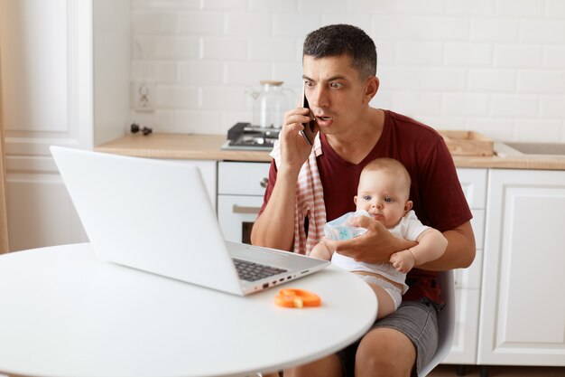 Schockiert, erstaunt, dunkelhaariger, gutaussehender Mann, der ein lässiges T-Shirt mit einem Handtuch auf der Schulter trägt, mit Laptop am Tisch sitzt, ein Baby in den Händen hält, telefoniert und auf die Anzeige des Notebooks schaut.