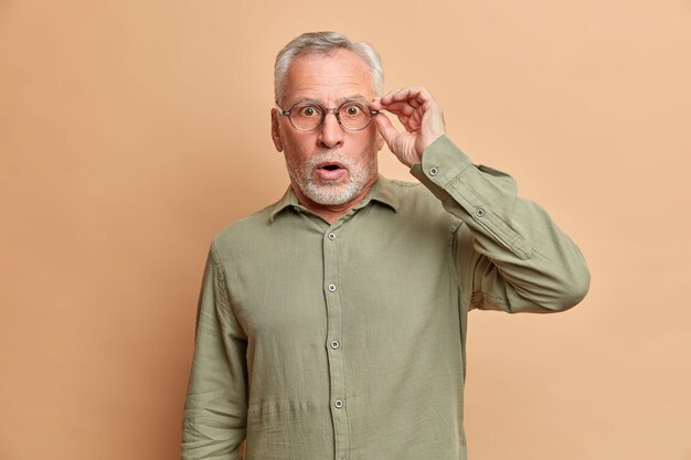 Schockiert gutaussehend betäubt bärtig älterer mann hat graues haar öffnet mund weit hält hand auf brille kann nicht an schockierende nachrichten glauben trägt formelle hemd posen gegen braune studiowand