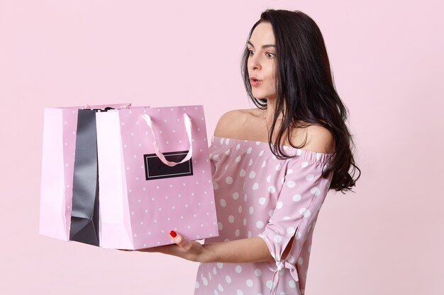 Schockiert überrascht europäische junge Frau hält Taschen, erstaunt, viele Geschenke zu erhalten, gekleidet in Tupfenkleid, will Geschenk öffnen, posiert auf rosa. Menschen und Einkaufskonzept