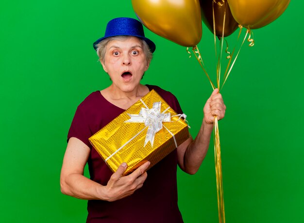 Schockierte ältere Frau, die Partyhut trägt, hält Heliumballons und Geschenkbox auf Grün