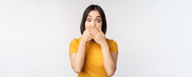 Schockierte asiatische Frau bedeckt den Mund mit Händen, die erschrocken aussehen, mit sprachlosem Gesichtsausdruck, der im gelben T-Shirt vor weißem Hintergrund steht