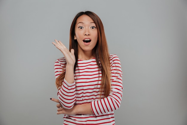 Schockierte asiatische Frau im Pullover, der die Kamera mit offenem Mund über grauem Hintergrund betrachtet