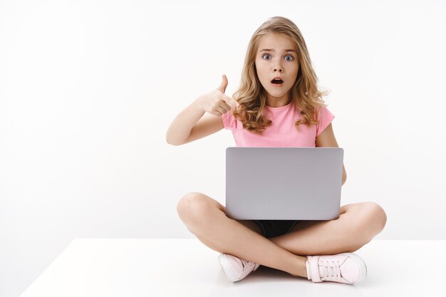 Schockierte besorgte süße blonde Mädchen kleine Tochter zeigt Mama gruseligen Videocomputer, sitzt überkreuzte Beine, hält den Laptop, zeigt auf den Gadget-Bildschirm, keucht verängstigt und nervös, starrt auf die Front verärgert, weiße Wand