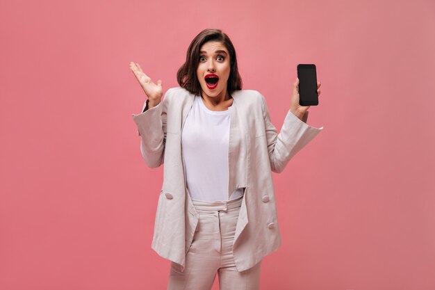 Schockierte Dame im Baumwollanzug demonstriert Telefon auf rosa Hintergrund. Überraschte Frau mit hellen Lippen in weißen Kleidern hält Telefon in ihren Händen.