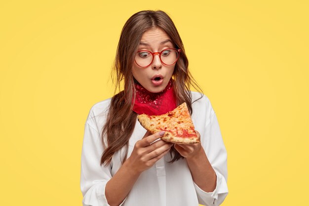 Schockierte emotionale Dame schaut mit Überraschung auf ein Stück Pizza, erstaunt über seinen wunderbaren Geschmack, trägt eine Brille und ein weißes Hemd, Modelle vor gelber Wand. Menschen-, Reaktions- und Ernährungskonzept