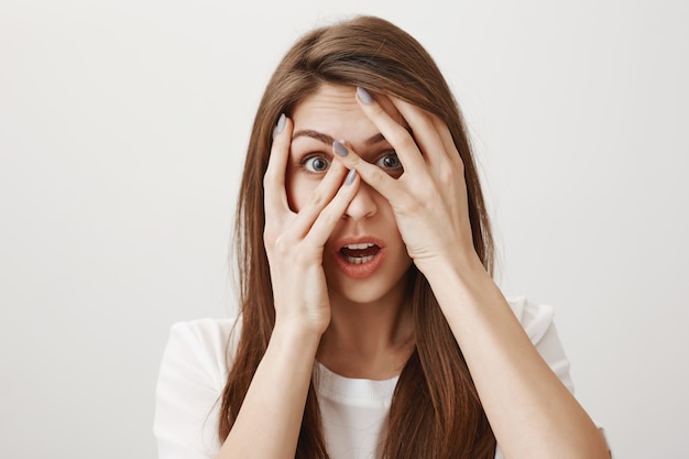 Schockierte Frau, die durch Finger mit erschrockenem Gesicht späht