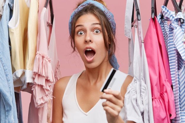 Schockierte Frau im weißen T-Shirt, in der Nähe des Kleiderständers stehend, Kreditkarte in der Hand, überrascht, dass sie kein Geld bei ihrem Buchhalter hat und ihre Garderobe auffrischen möchte. Outfit kaufen