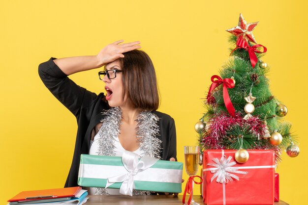 Schockierte Geschäftsdame im Anzug mit Brille, die ihr Geschenk zeigt und an einem Tisch mit einem Weihnachtsbaum darauf im Büro sitzt