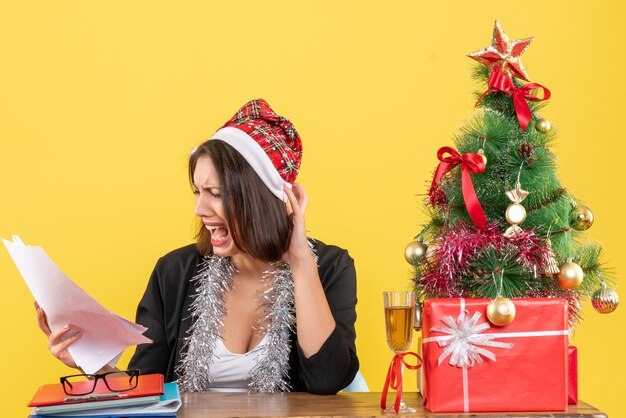 Schockierte Geschäftsdame im Anzug mit Weihnachtsmannhut und Neujahrsdekorationen, die Dokumente halten und an einem Tisch mit einem Weihnachtsbaum darauf im Büro sitzen