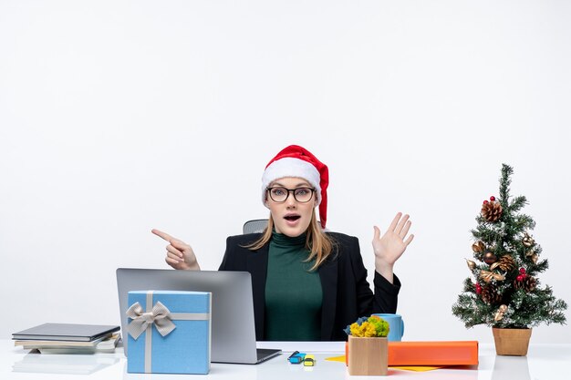 Schockierte Geschäftsfrau mit Weihnachtsmannhut, die an einem Tisch mit einem Weihnachtsbaum und einem Geschenk darauf sitzt und sich auf etwas sorgfältig auf weißem Hintergrund konzentriert