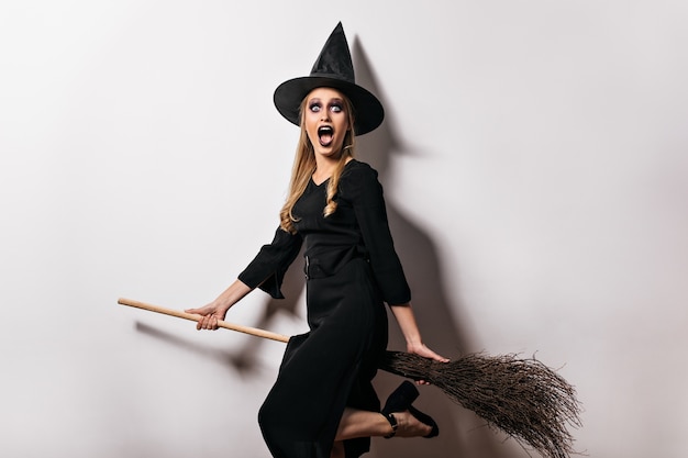 Kostenloses Foto schockierte hexe mit schwarzem make-up, das in halloween aufwirft. charmante blonde dame im magischen hut, der besen hält.