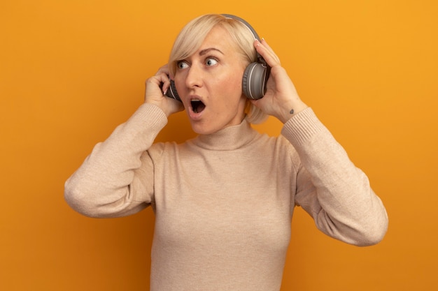 Kostenloses Foto schockierte hübsche blonde slawische frau auf kopfhörern schaut seite auf orange