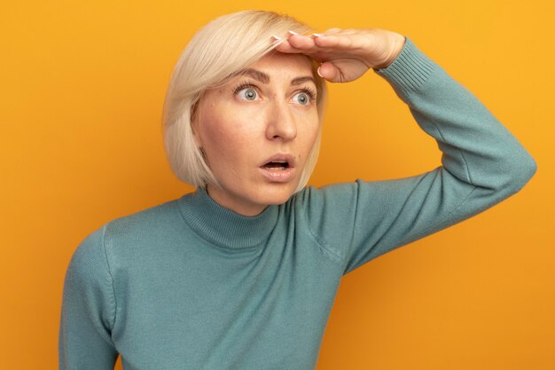 Schockierte hübsche blonde slawische Frau hält Handfläche an der Stirn und betrachtet Seite isoliert auf orange Wand
