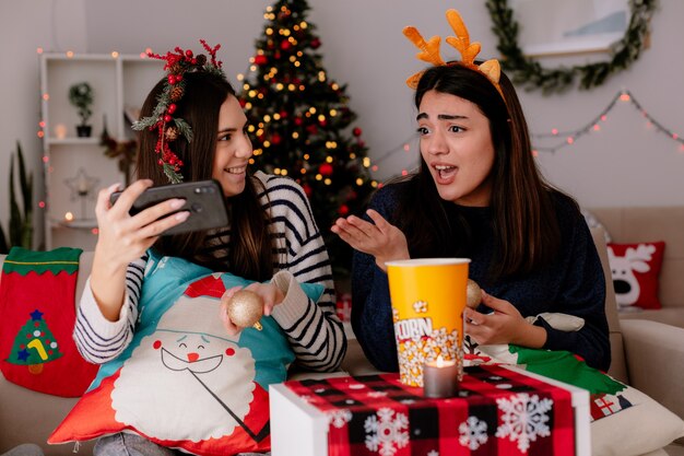 Schockierte hübsche junge Mädchen mit Stechpalmenkranz und Rentierstirnband schauen auf das Telefon, das auf Sesseln sitzt und die Weihnachtszeit zu Hause genießt