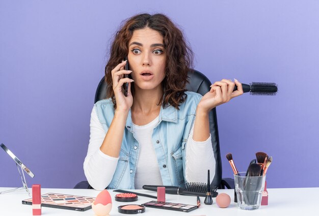 Schockierte hübsche kaukasische Frau, die am Tisch mit Make-up-Tools sitzt, die am Telefon spricht und den Kamm isoliert auf lila Wand mit Kopienraum hält?