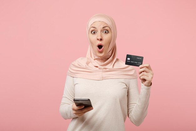 Schockierte junge arabische muslimische frau in hijab-leichter kleidung posiert isoliert auf rosa hintergrund. menschen religiösen islam lifestyle-konzept. mock-up-kopienbereich. verwenden sie das mobiltelefon und halten sie eine kreditkarte bereit.