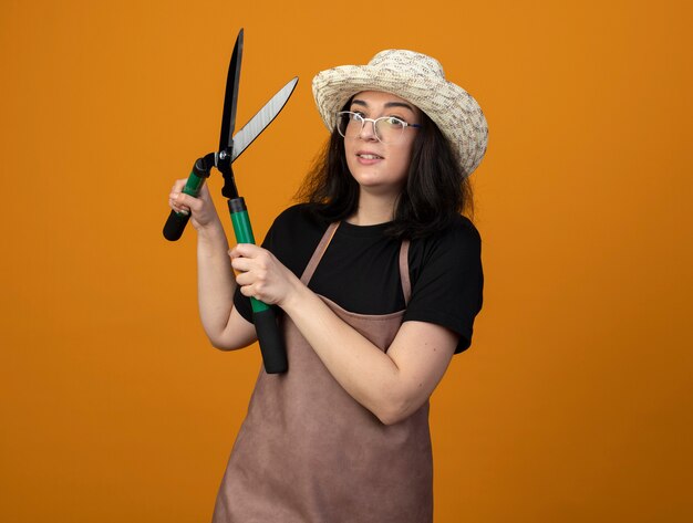 Schockierte junge brünette Gärtnerin in optischen Gläsern und in Uniform mit Gartenhut hält Gartenschere isoliert auf orange Wand