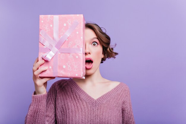 Schockierte junge Frau, die rosa Schachtel mit Band verziert hält. Innenporträt des kurzhaarigen Mädchens im Pullover, der mit Geburtstagsgeschenk auf lila Wand aufwirft.
