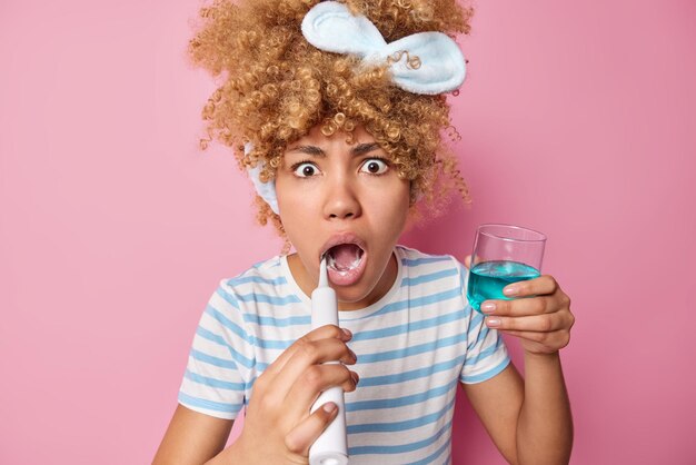 Schockierte junge Frau mit lockigem Haar trägt ein Stirnband und ein lässig gestreiftes T-Shirt bürstet die Zähne mit einer elektrischen Bürste und hält ein Glas blaues Mundwasser isoliert über rosa Hintergrund Tägliches Hygienekonzept