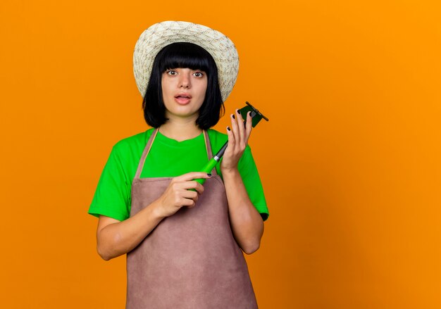 Schockierte junge Gärtnerin in Uniform mit Gartenhut hält Hacke Rechen