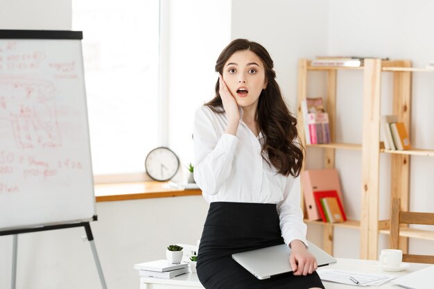 Schockierte junge Geschäftsfrau, die mit etwas vor dem Sitzen im modernen Büro überrascht war