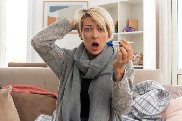 Schockierte junge kranke slawische Frau mit Schal um den Hals, die die Hand auf den Kopf legt und das Thermometer auf der Couch im Wohnzimmer hält