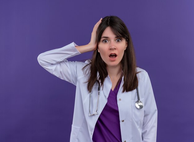 Schockierte junge Ärztin im medizinischen Gewand mit Stethoskop legt Hand auf Kopf und schaut auf Kamera auf lokalisiertem violettem Hintergrund