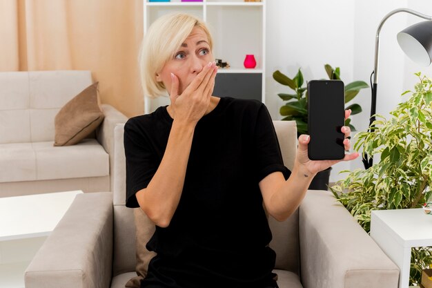 Schockierte schöne blonde russische Frau sitzt auf Sessel, der Hand auf Mund hält Telefon im Wohnzimmer hält