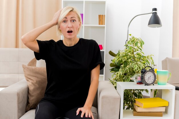 Schockierte schöne blonde russische Frau sitzt auf Sessel, der Kopf im Wohnzimmer hält